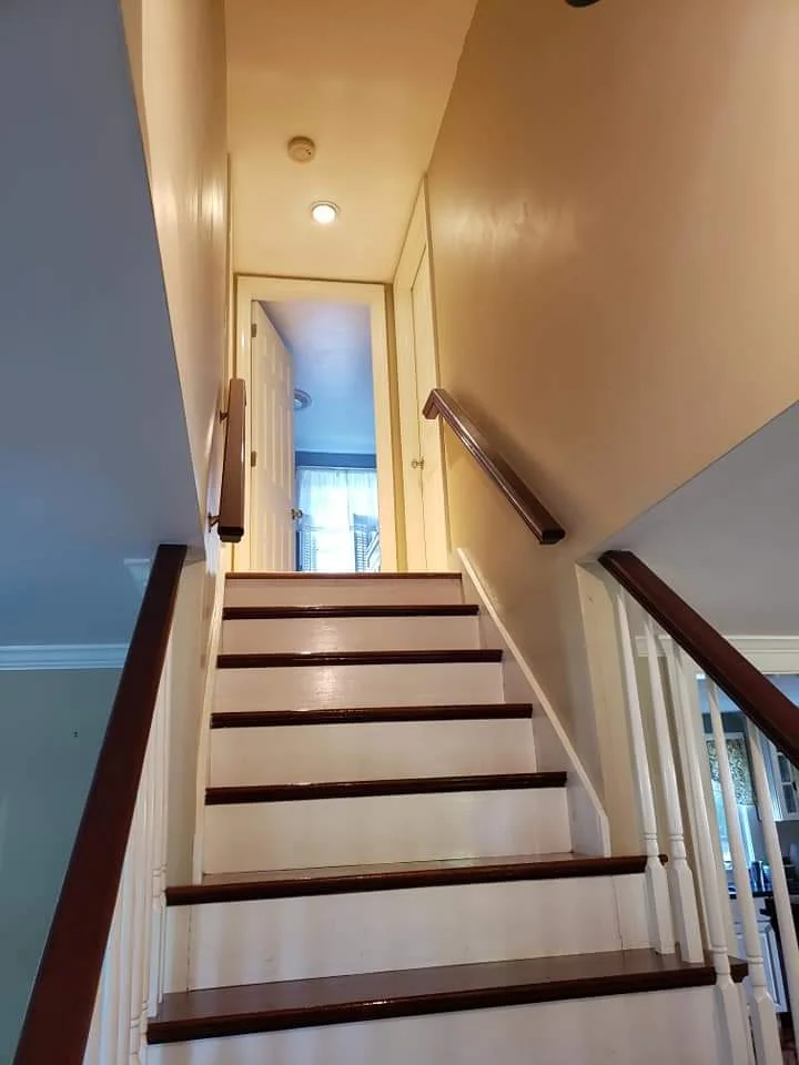 Before: Stairwell with white painted stair faces, stained wood treads, and stained wood railing. The walls are unpainted, and the banister is white. Flagler County Painter, Residential Painting, Stairwell Transformation, T&B Paint Pros LLC.