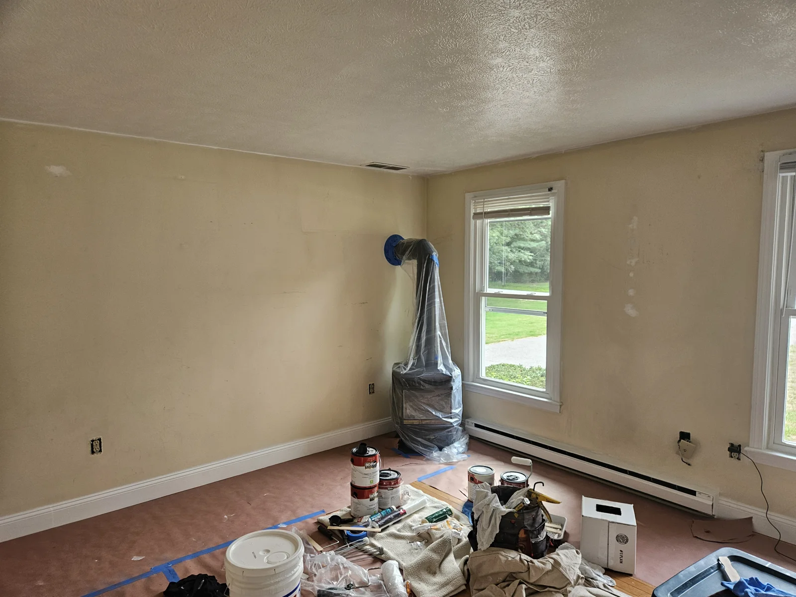 Living room with yellow walls and wood stove wrapped in plastic for protection. Floors covered as part of the prep process for repainting. T&B Paint Pros LLC, Flagler County Painter.