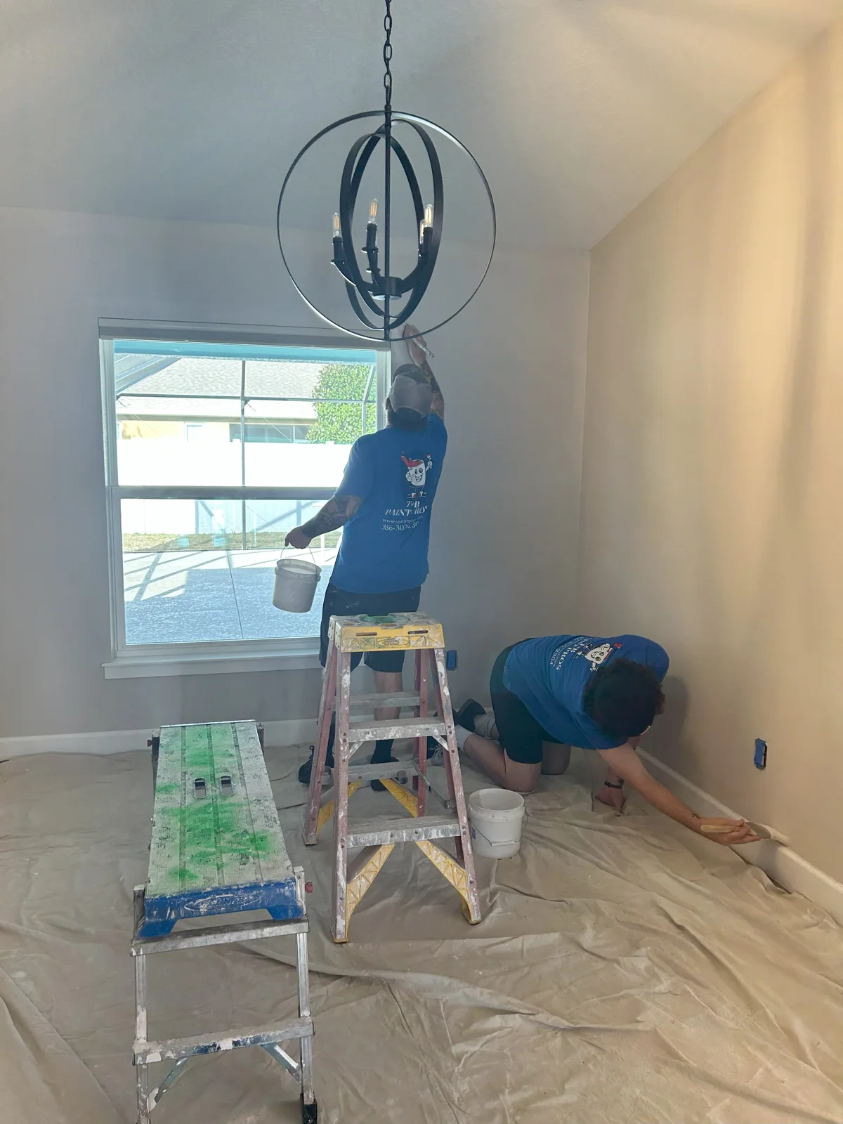 Beau and Tim painting the dining area in Palm Coast, FL. Tim is on the floor painting the trim, while Beau reaches up to coat the walls in Sherwin Williams Popular Gray. Floors are covered for protection. T&B Paint Pros LLC in action.